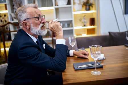 Senior grey-haired man holding and smoke e-cigarette in rich restaurant . IQOS, modern technologies, stop smokeの写真素材
