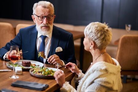 Pretty lady and elderly grey haired man in tuxedo having friendly conversation in restaurant. Colleagues after work discuss and eatの写真素材