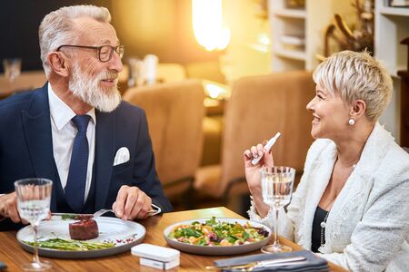 Elderly couple have dinner in rich restaurant while using IQOS, e-cigarette. Handsome man in tuxedo smile, look at woman and speak.の写真素材