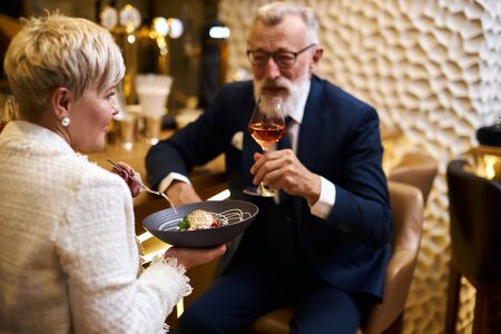 Mature couple of caucasian man and woman sit in restaurant and eat dessert, drink glass of wine. Male in tuxedo, female in white blazer. Woman holding dishの写真素材