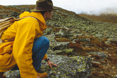 Young man in yellow raincoat and grren nat is sitting on cliffs edge and looking to misty valley...