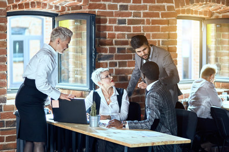 Four people dressed in stylish office clothes discuss task while sit at table. Window open. Behind...