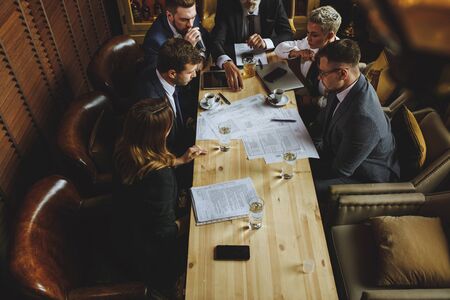Determined business partners actively studying business documents at wooden conference table. Busy young man with a beard holds glass of water, inspect paper files, plan viewの写真素材