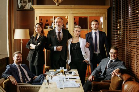Elderly man embracing blonde woman in a white jacket, brunette standing behind with arms crossed, young red-bearded guy smiles broadly sitting in brown chair,man in glasses looks joyfully at cameraの写真素材