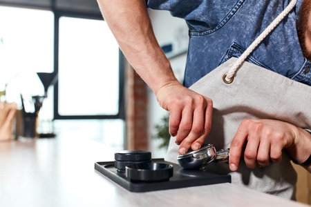 Hands of bartender pressing ground coffee in tamper, making hot fresh espresso behind light coloured...