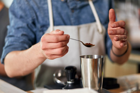 Hands of male beared barista, keep little spoon with ground coffee over metal pot, measure coffee...