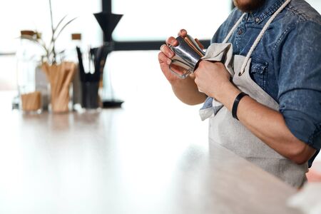 Hands of solid man wearing white neat apron holding towel, cleaning steel pitcher in brightly lighted room, background of window in modern restaurantの写真素材