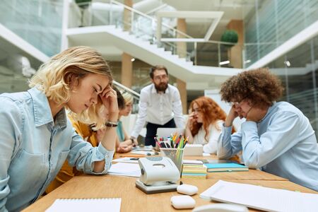 Serious concentrated young female designer, sits at light coloured table, thoughtfully stares at papers, puts elbow on desk , keeps head with delicate hand, brainstorming conceptの写真素材