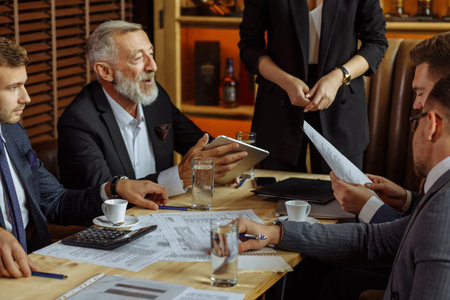 businessmen in stylish suits sit at table in cozy office discussing strategic plans for future...