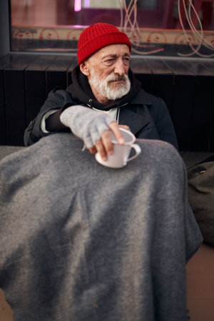 Homeless man begging in the center of city, sitting on the floor next to modern cafe and looking...