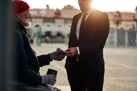 Homeless senior man hold out his hand to get money from unrecognizable rich man. Street lifeの写真素材