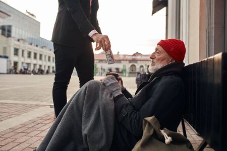 Rich man came up and help homeless man sitting on the floor. Help, money donation to vagrant male in the streetの写真素材