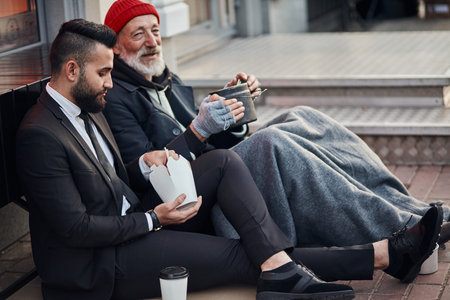Handsome businessman in suit sitting on floor with homeless man together, listen to his story of life. Contrast people, rich and poor, but doesnt matterの写真素材