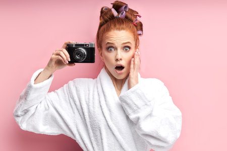 Open-eyed young girl in bathrobe and curlers in hair taking photo with camera.