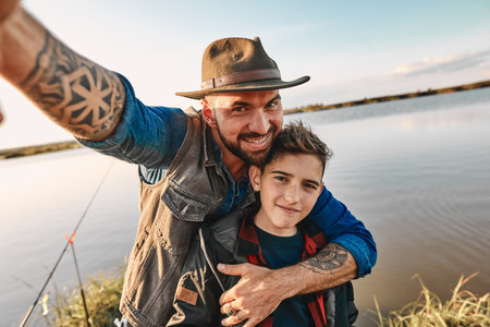 Father hugs son and takes selfie, which will stand on table at home. They have fun. Background lake.