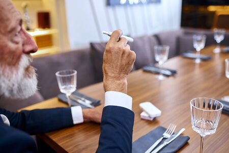 Portrait senior bearded man grey haired in suite sit in restaurant looking electronic cigarette. Modern technologies.の写真素材