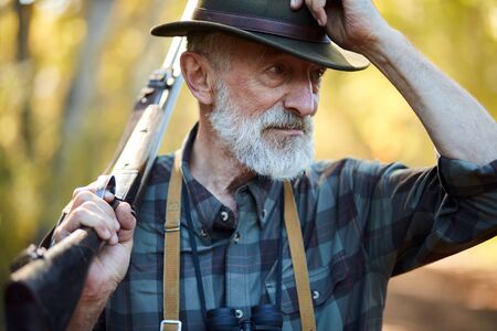 Senior hunter on birds holding gun on shoulder, straighten hat, looking away. Forest backgroundの写真素材