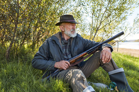 Bearded male with shotgun sitting on grass, have rest after hunting, wearing hat, coat and rubber bootsの写真素材