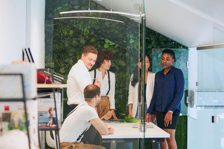 young businesspeople in office boardroom gathered together around white table, discussing their...