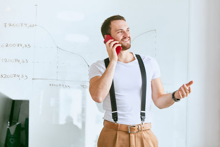 Young businessman wearing white t-shirt and trousers with suspenders look side, talking on phone...