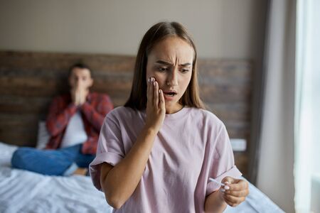 Frustrated sad female standing in bedroom, staring at pregnancy test with opened mouth, raising hand towards cheek with surprised expression, demonstrating shock and sadness, close upの写真素材
