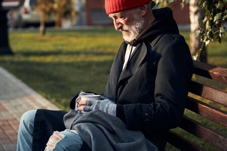 Mature man without shelter sitting in street clothes on bench, without food and money. Drooping man lowered his head down. Side view on homeless person in coat and red capの写真素材