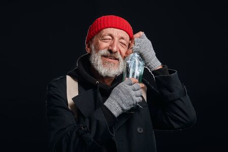 Happy old beardy man wearing red hat and black coat happily hugging bottle of drinking. Isolated over black backgroundの写真素材