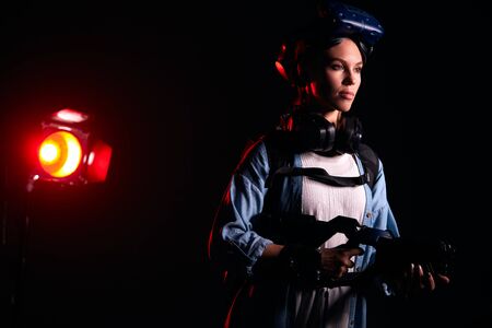 Confident female stand posing with VR weapon, headsets, goggles on head. Isolated studio photo. Light red smoky backgroundの写真素材