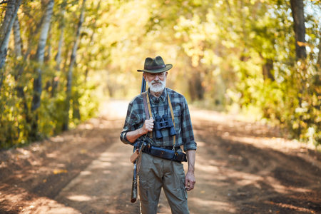 Portrait of caucasian bearded hunter with grey beard stand in autumn forest looking at camera....