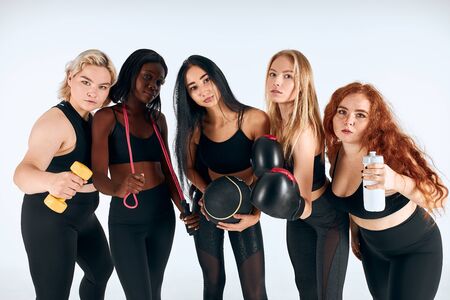 Sportive girls in sport wearing stand together with boxing gloves, dumbbells and other sportive things, look at camera isolated, white backgroundの写真素材