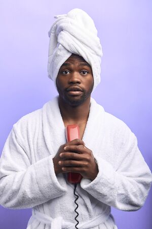 Shocked man. Susprised guy in towel and bathrobe after shower stand looking at camera with opened mouth isolated over purple backgroundの写真素材