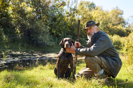 Good-looking senior man satisfied with hunting on lake, sit next to his dog, in forest, sunny day