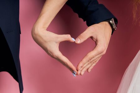 Portrait closeup hands of couple in love forming hear isolated over pink background. man in tuxedoの写真素材