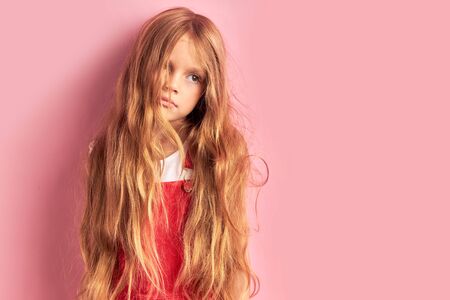 Portrait of shaggy caucasian girl in red overalls with long golden hair looking side with sadness in eyes, after sleeping. Pink backgroundの写真素材