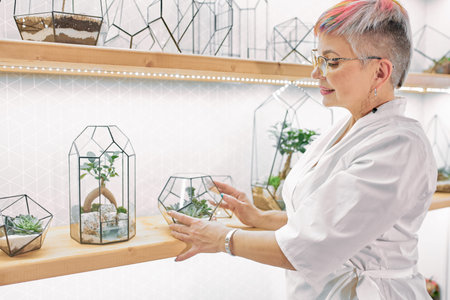 Elderly woman florist look after plants in office, work place. Woman wearing white suit in office full of plants. Light roomの写真素材