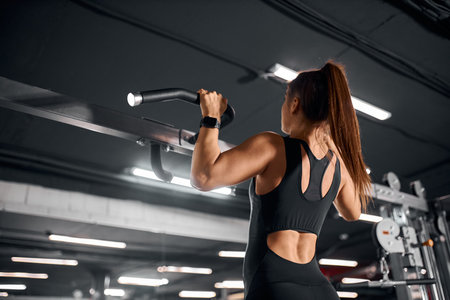 Young strong female powerlifter dressed in black sexy sportswear, pulling handles of fitness machine...