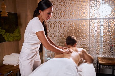 Young pretty woman with long brunette hair, dressed in white uniform doing professional tissue massage to male client in medical clinic, satisfaction and pleasure concept, indoor shotの写真素材