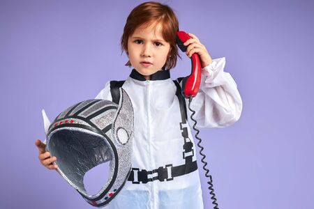 Little spaceman kid speaking on landline phone, dressed in white suit and holding helmet, isolated over purple backgroundの写真素材