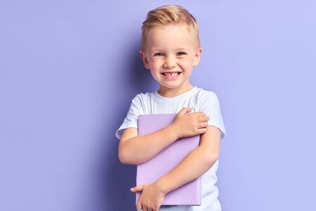 Portrait of blond kid boy holding book happily looking at camera , purple background. Kid involved in educationの写真素材