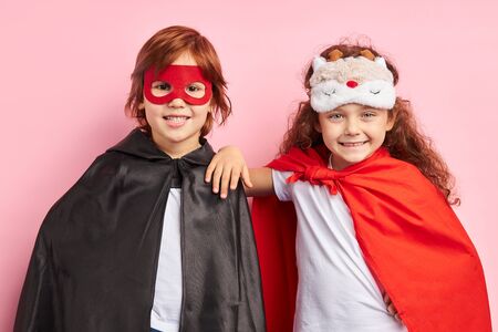 Two cheerful children wearing cloaks and mask, imagine themselves as heroes. Portrait, isolated over pink backgroundの写真素材