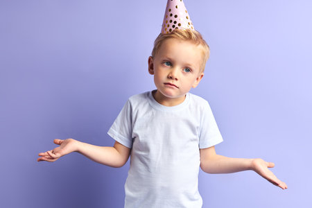 Portrait of upset boy wearing birthday cap stand spreading arms in different directions, isolated...