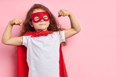 Beautiful little curly girl wearing red hero suit and mask showing how she is strong isolated over pink backgroundの写真素材