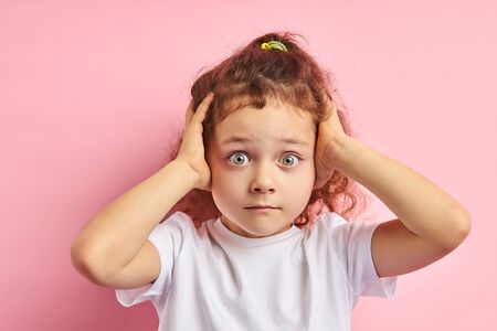 Bright emotions of little caucasian girl with auburn hair, clutching head while standing on pink backgroundの写真素材