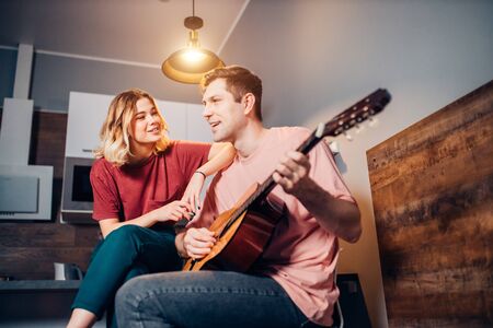 caucasian young man wearing casual clothes sit with her blond short-haired girlfriend playing guitar for her and singingの写真素材