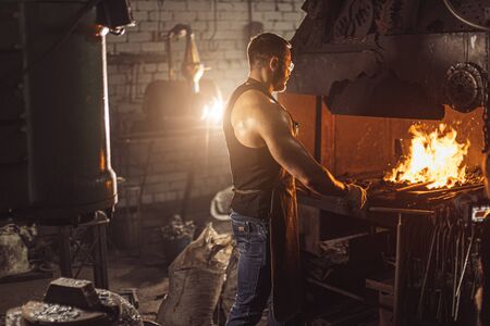 Young caucasian man blacksmith, strong iron worker in workshop, work place, isolated dark space with furnaceの写真素材