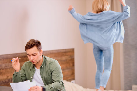 tired busy man sit on table working on laptop, checking documents while his wife jumping on bed, in...