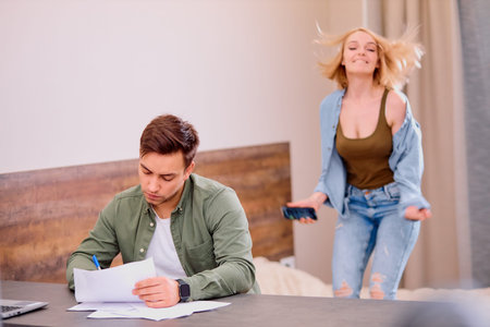 busy tired man sit working at home while his woman wife or girlfriend jump, smile in the background,...