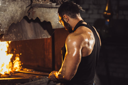 young caucasian powerful blacksmith man with muscular hands, stand opposite of fire, heating steel...