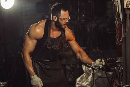 brutal strong caucasian man work as forger blacksmith and making metal in workshop, dressed in dark leather apron on naked bodyの写真素材