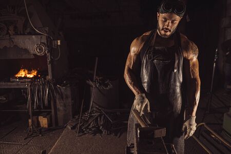 confident good-lloking artisan in leather brown apron or uniform, young caucasian forger with beard wearing glovesの写真素材
